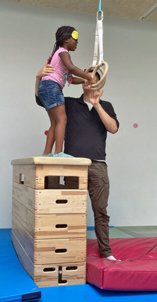 Ein Erzieher hilt einem Kind das mit verbundenen Augen von einem Kasten auf eine weiche Matte springt