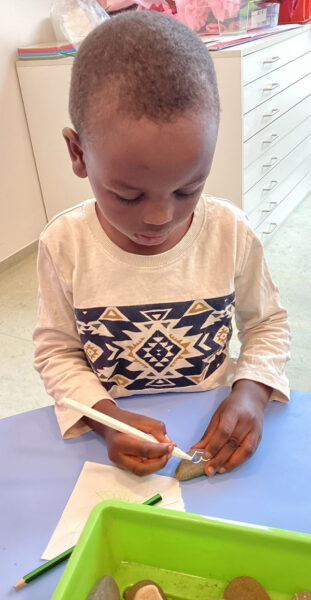 Ein Kind sitzt am Tisch und bemalt eine Stein mit einem weiss-bunten Herz